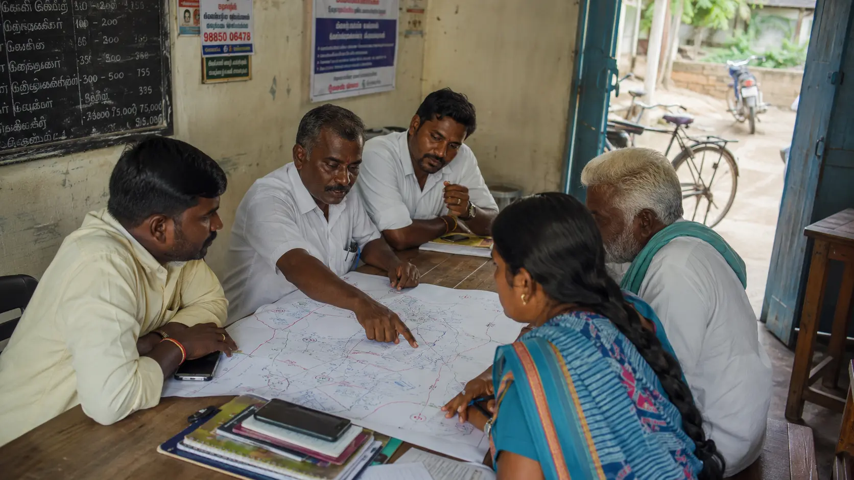 Small campaign team discussing strategy around a hand-drawn ward map in a village office